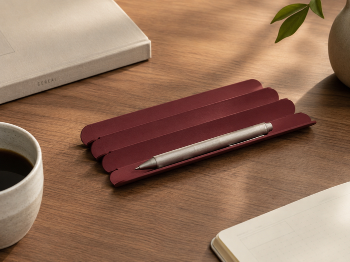 A burgundy aluminum pen stand holding a silver pen, styled on a wooden desk with a coffee cup, notebook, and book nearby.